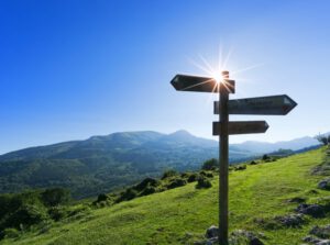 Ein Bergpfad, auf dem ein Wegweiser in 3 Richtungen zeigt. Im Hintergrund die Sonne. Stehlt für berufliche Neuorientierung durch Coaching.