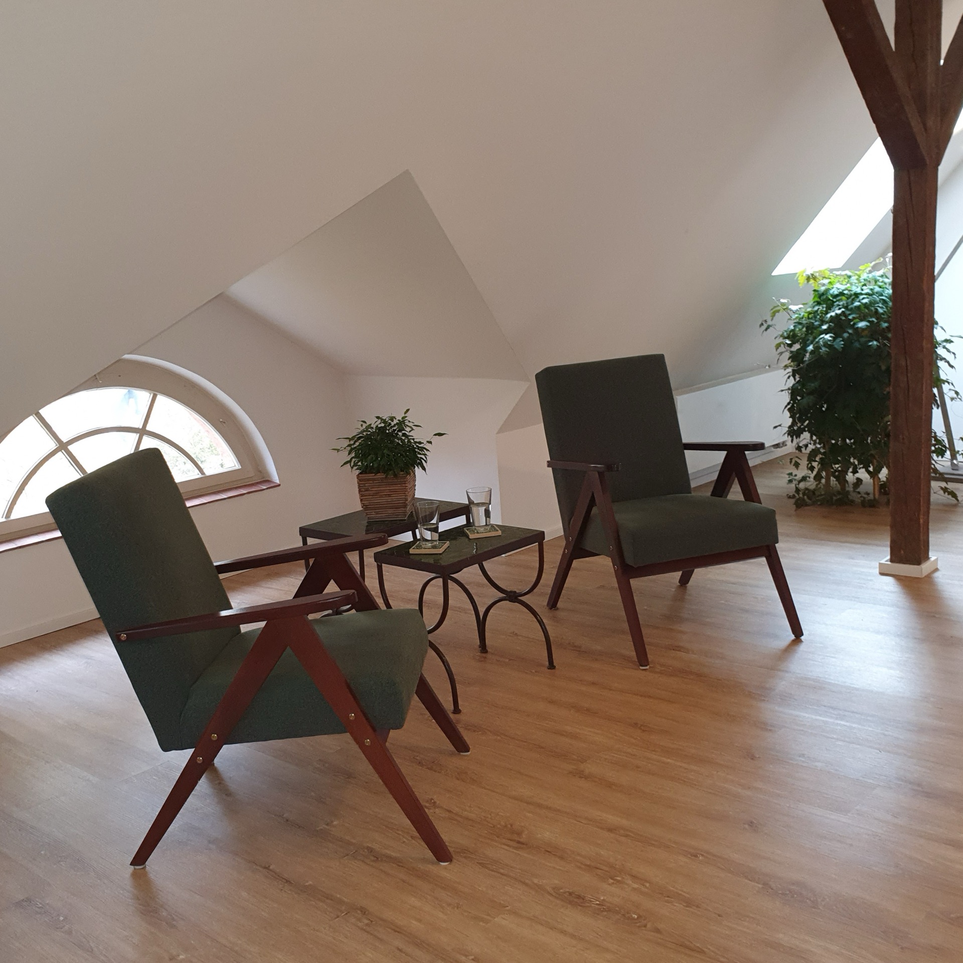 Zwei Sessel, zwei Tischchen. Pflanzen. Ein Raum mit Dachschräge und hellem Boden