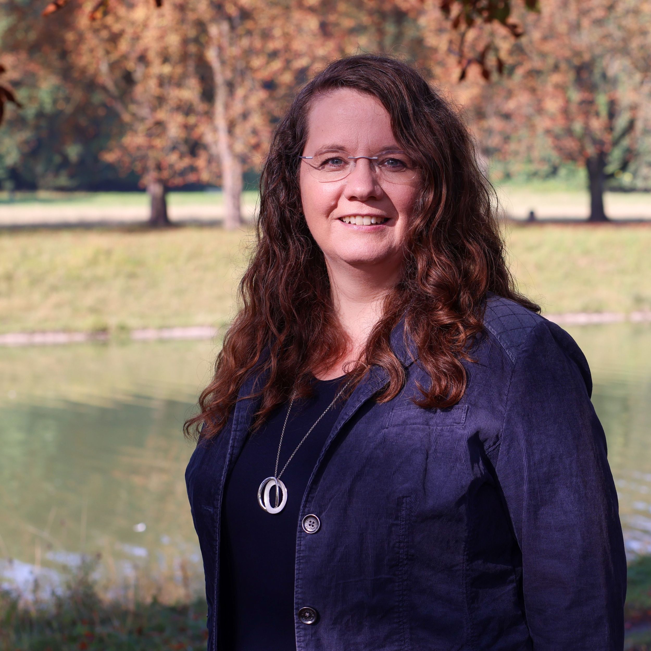 Katharina Temme steht vor einem Gewässer. Im Hintergrund herbstliche Bäume.