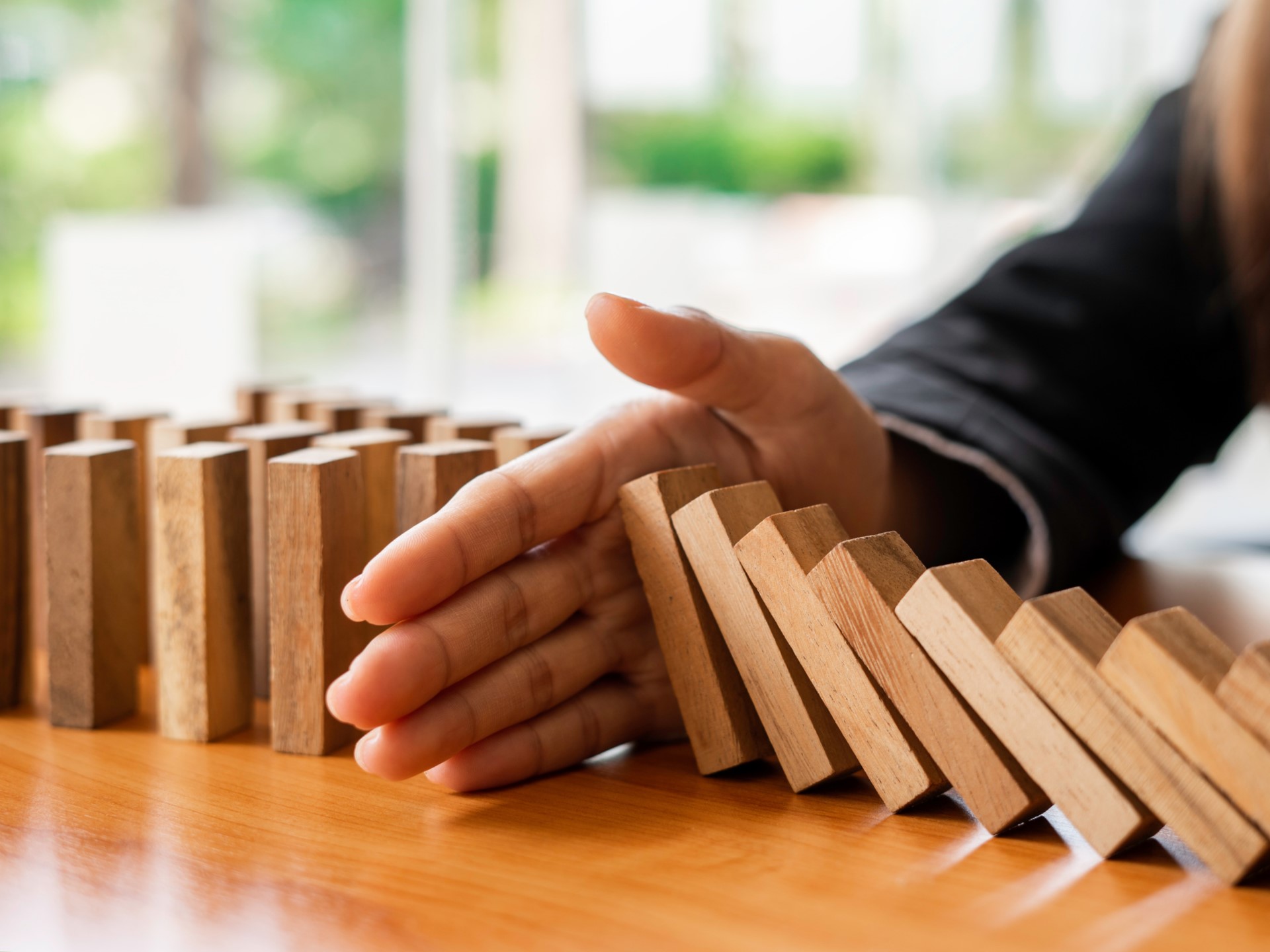 Auf einem Tisch sind Holzsteine wie beim Domino platziert. Eine Hand unterbricht das Umfallen.