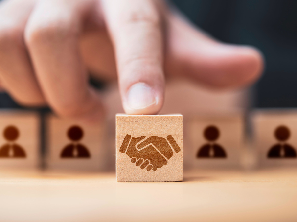 Ein Würfel, auf dem ein Handschütteln zu sehen ist, wird in die Mitte des Tischs geschoben und symbolisiert Streitschlichtung durch Mediation.